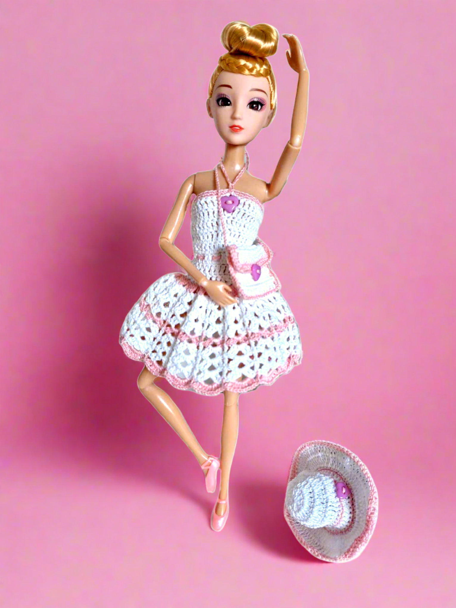 Vista frontal da boneca com o seu bonito vestido curto, justo na parte de cima e uma saia tipo balão. Este é maioritariamente branco com a exceção de alguns pormenores como a fita a volta do pescoço, usada para prender o vestido, um pequeno botão em forma de coração na parte de peito e três riscas na saia. Este conjunto é ainda composto por um bonito chapéu redondo  e uma pequena mala a tiracolo. Tal como o vestido este dois acessórios também são brancos com pequenos pormenores em cor de rosa.       