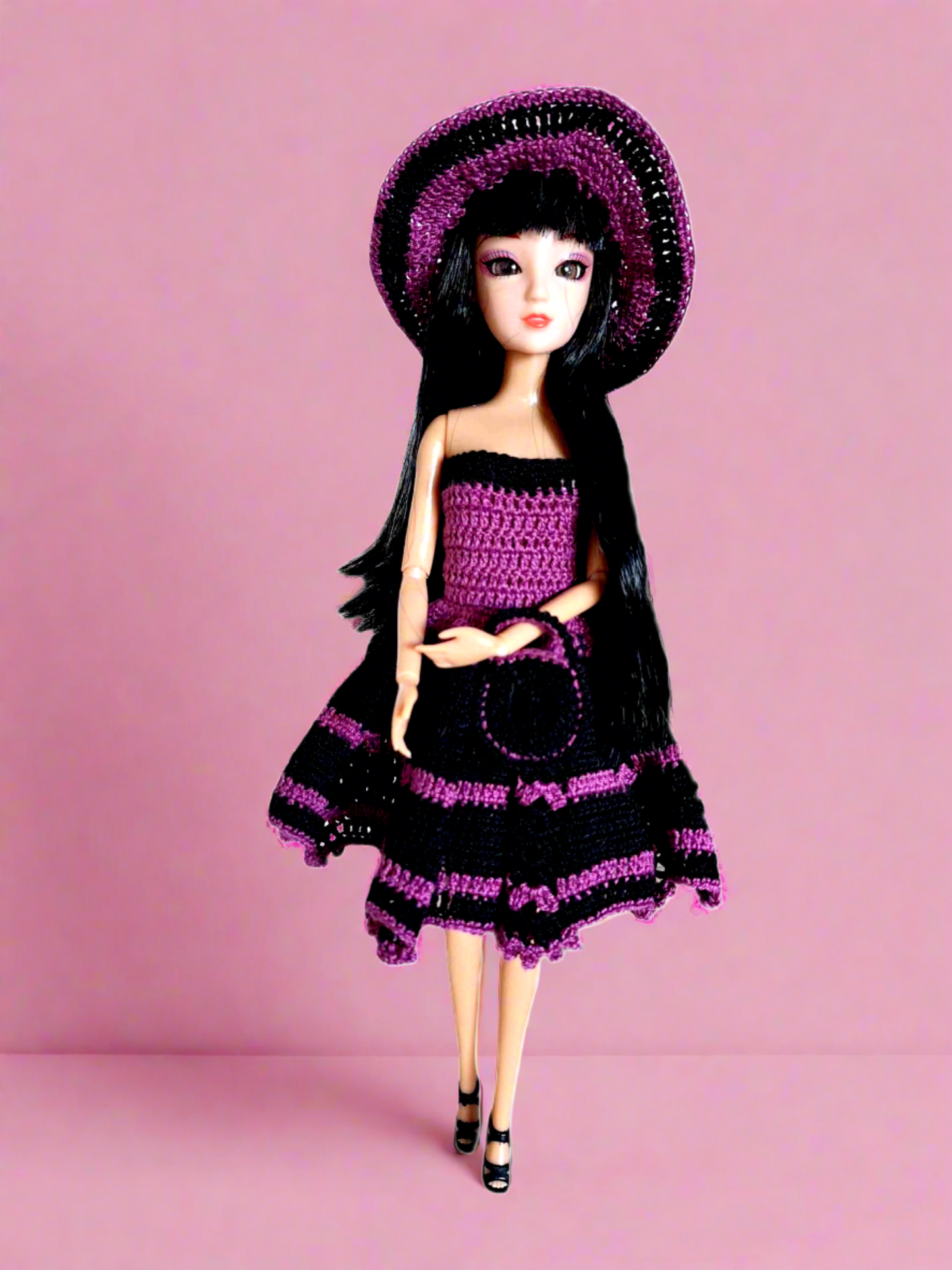 Vista frontal da boneca, com o seu vestido de cocktail, sem alças, com a parte de cima justa e uma saia mais larga com o comprimento pelo meio da perna. Tem ainda como acessórios um bonito chapéu e uma pequena bolsa. Este conjunto é todo em roxo e preto dando-lhe um ar muito requintado.  