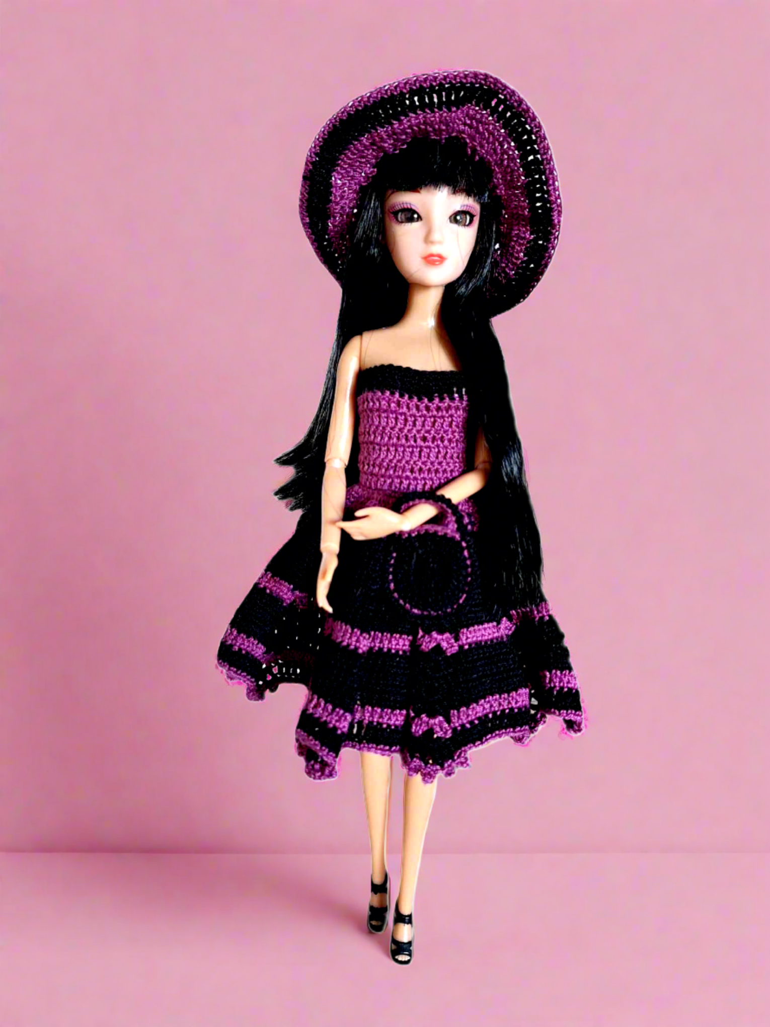 Vista frontal da boneca, com o seu vestido de cocktail, sem alças, com a parte de cima justa e uma saia mais larga com o comprimento pelo meio da perna. Tem ainda como acessórios um bonito chapéu e uma pequena bolsa. Este conjunto é todo em roxo e preto dando-lhe um ar muito requintado.  