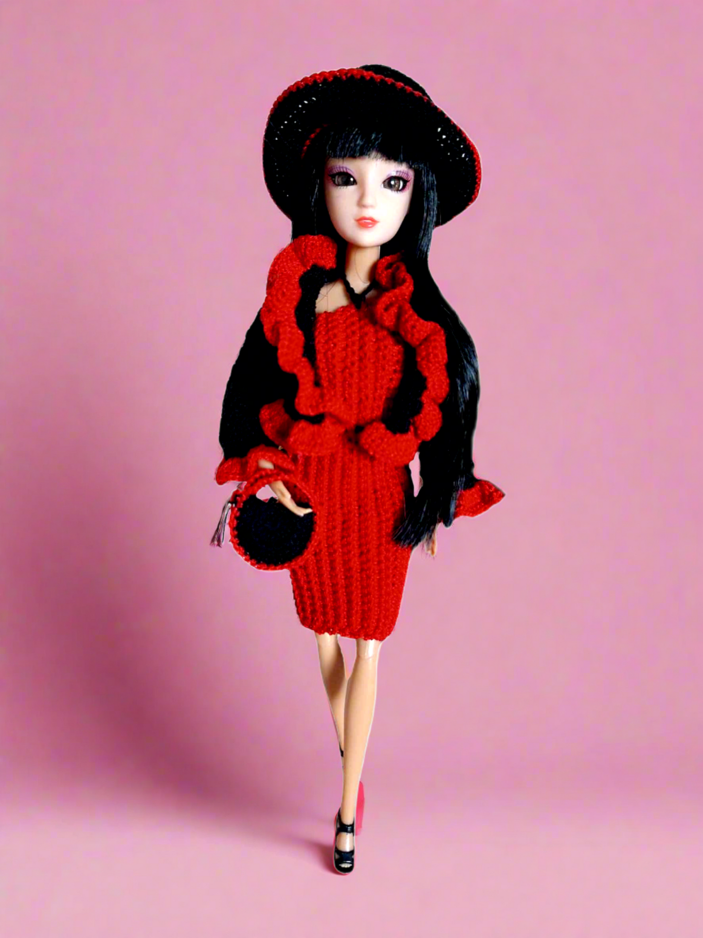 Vista frontal da boneca, com o seu vestido justo e completamente vermelho, casaco preto com folhos nos punhos e parte da frente em vermelho, chapéu e bolsa, ambos redondos com pequenos pormenores em vermelho.