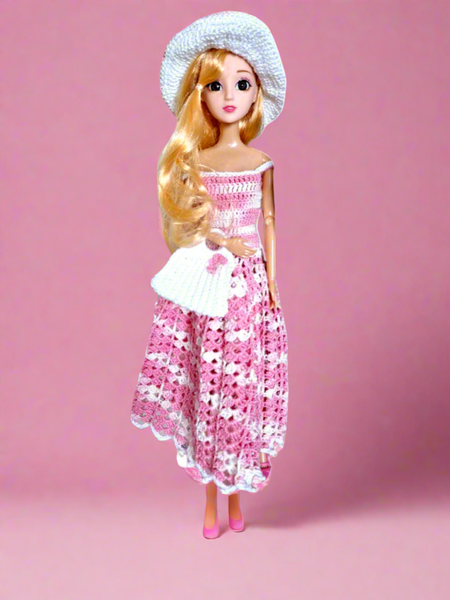 Vista frontal do conjunto. Vestido comprido em rosa matizado, com pequenas alças que caem dos ombros. É de destacar ainda a bonita mala de mão e um chapéu, ambos em branco com pequenos pormenores em rosa. A mala tem uma pequena flor na parte frontal e o chapéu tem um elegante laço em cetim.