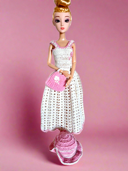 Vista frontal do conjunto, composto por um longo vestido pérola com uma pequena flor na zona do peito da cor rosa. Tem ainda um chapéu e uma bonita mala de mão, ambos rosa com alguns pormenores em branco, como a pequena flor e as alças da mala.