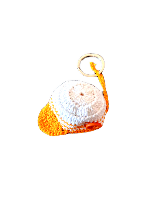 Porta chaves com o formato de um pequeno boné. Este é branco no centro e laranja no rebordo, pala e ainda num pequeno botão que adorna um dos lados deste item.