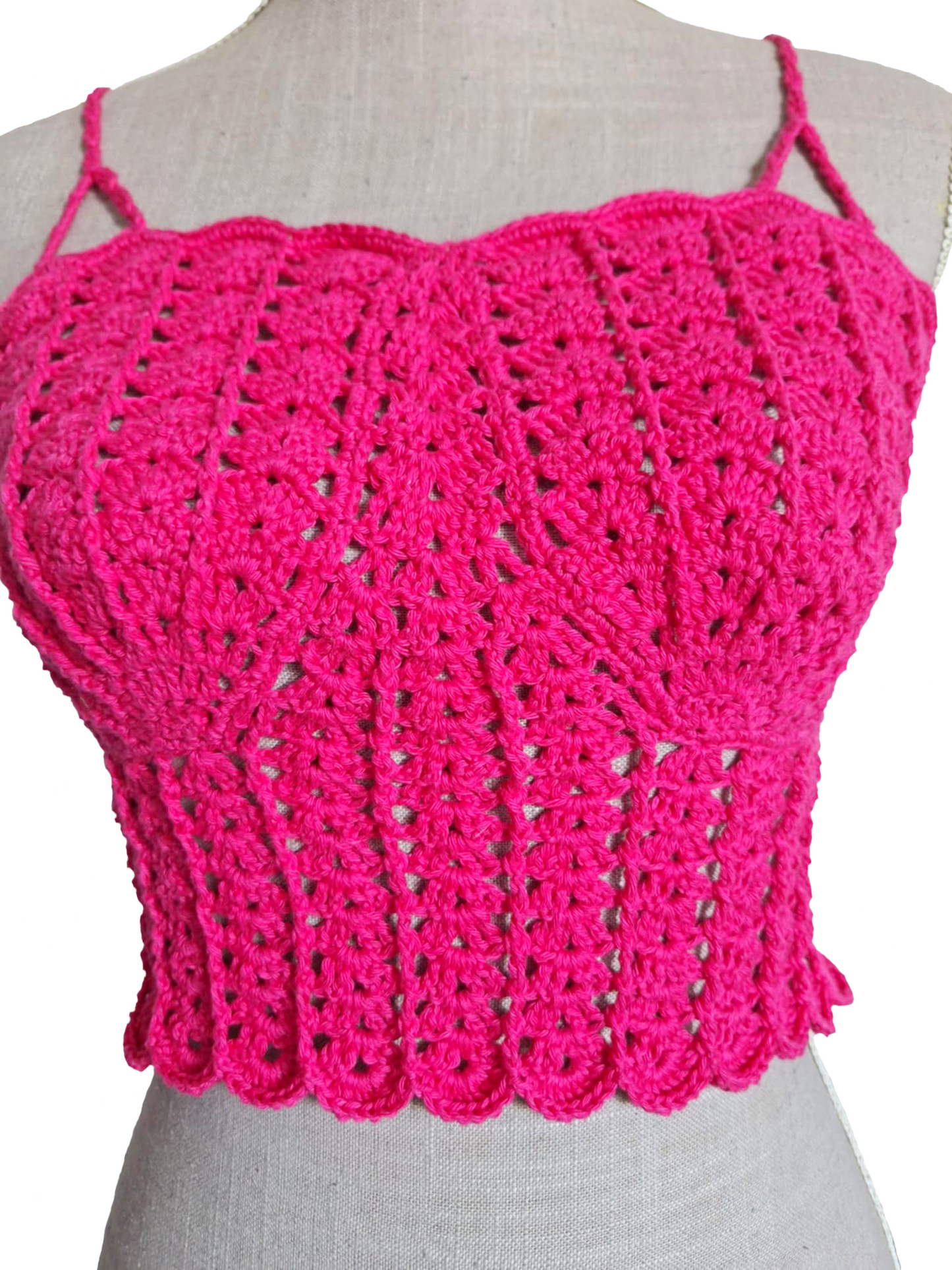 Imagem muito aproximada da parte frontal de um top de crochet fúcsia num manequim. A fotografia realça os pormenores do trabalho artesanal, com foco nos pontos em relevo, na textura das copas em forma de conchas e na base do top.