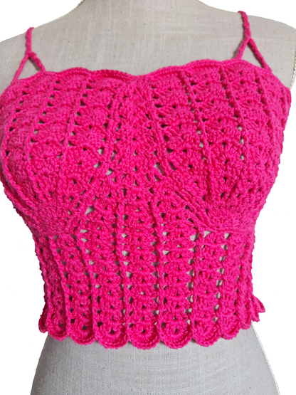 Imagem muito aproximada da parte frontal de um top de crochet fúcsia num manequim. A fotografia realça os pormenores do trabalho artesanal, com foco nos pontos em relevo, na textura das copas em forma de conchas e na base do top.