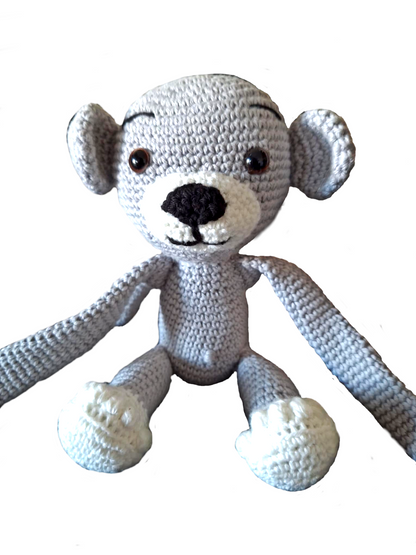 Macaquinho de crochet cinzento sentado de frente, com patas e focinho brancos. A imagem está aproximada, mostrando em detalhe a textura do crochet, os olhos castanhos de segurança, o nariz preto e a expressão amigável. Os braços e pernas longos estão estendidos à frente do corpo.