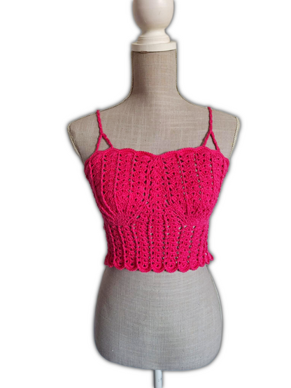 Top de crochet fúcsia em manequim, visto de frente. O modelo é cropped, com copas em forma de conchas, alças finas que sobem pelos ombros e design aberto abaixo do busto. A cor vibrante e o estilo destacam-se contra o fundo neutro.