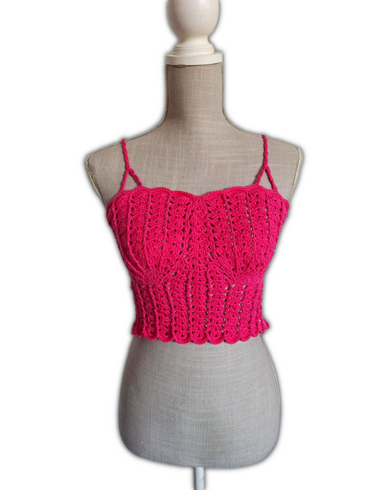 Top de crochet fúcsia em manequim, visto de frente. O modelo é cropped, com copas em forma de conchas, alças finas que sobem pelos ombros e design aberto abaixo do busto. A cor vibrante e o estilo destacam-se contra o fundo neutro.