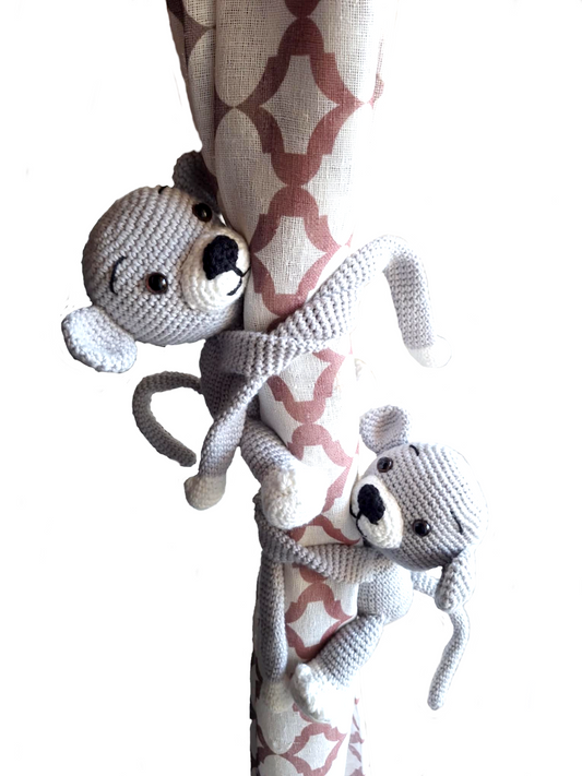 Dois macaquinhos de crochet cinzentos com patas brancas abraçam uma cortina. Têm olhos castanhos de segurança, nariz preto e expressão amigável. Feitos à mão com fio de algodão, com braços e pernas compridos que se enrolam à volta da cortina.