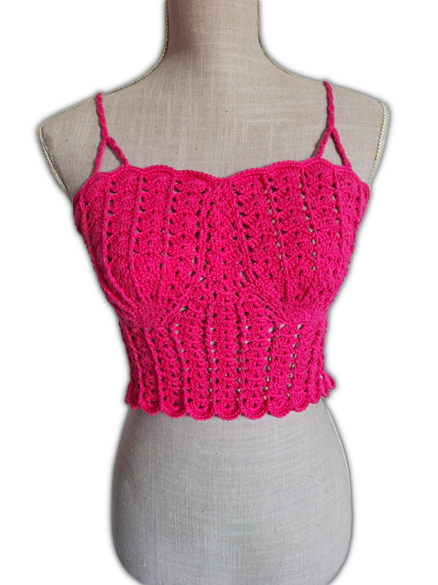 Close-up frontal de um top de crochet fúcsia num manequim. A imagem destaca as copas em forma de conchas e o trabalho detalhado dos pontos. A textura do crochet e o design do busto são claramente visíveis.