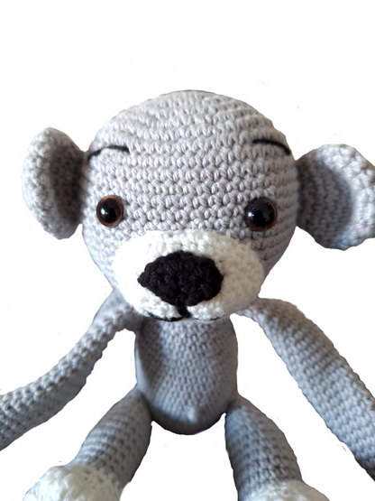 Close-up do rosto de um macaquinho de crochet cinzento. Mostra em detalhe os olhos castanhos de segurança, o nariz preto bordado, o focinho branco e a expressão simpática. A textura do crochet é visível, com pontos pequenos e uniformes.