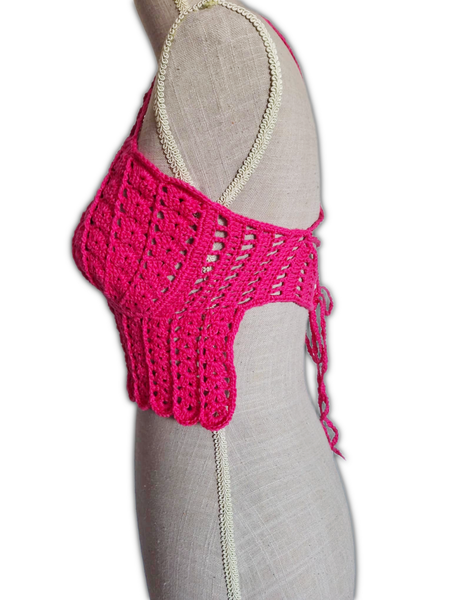 Vista lateral de um top de crochet fúcsia num manequim. A imagem mostra o perfil do busto com copas em forma de conchas, as alças finas sobre o ombro e parte da lateral aberta do top, destacando o caimento e a estrutura leve da peça.