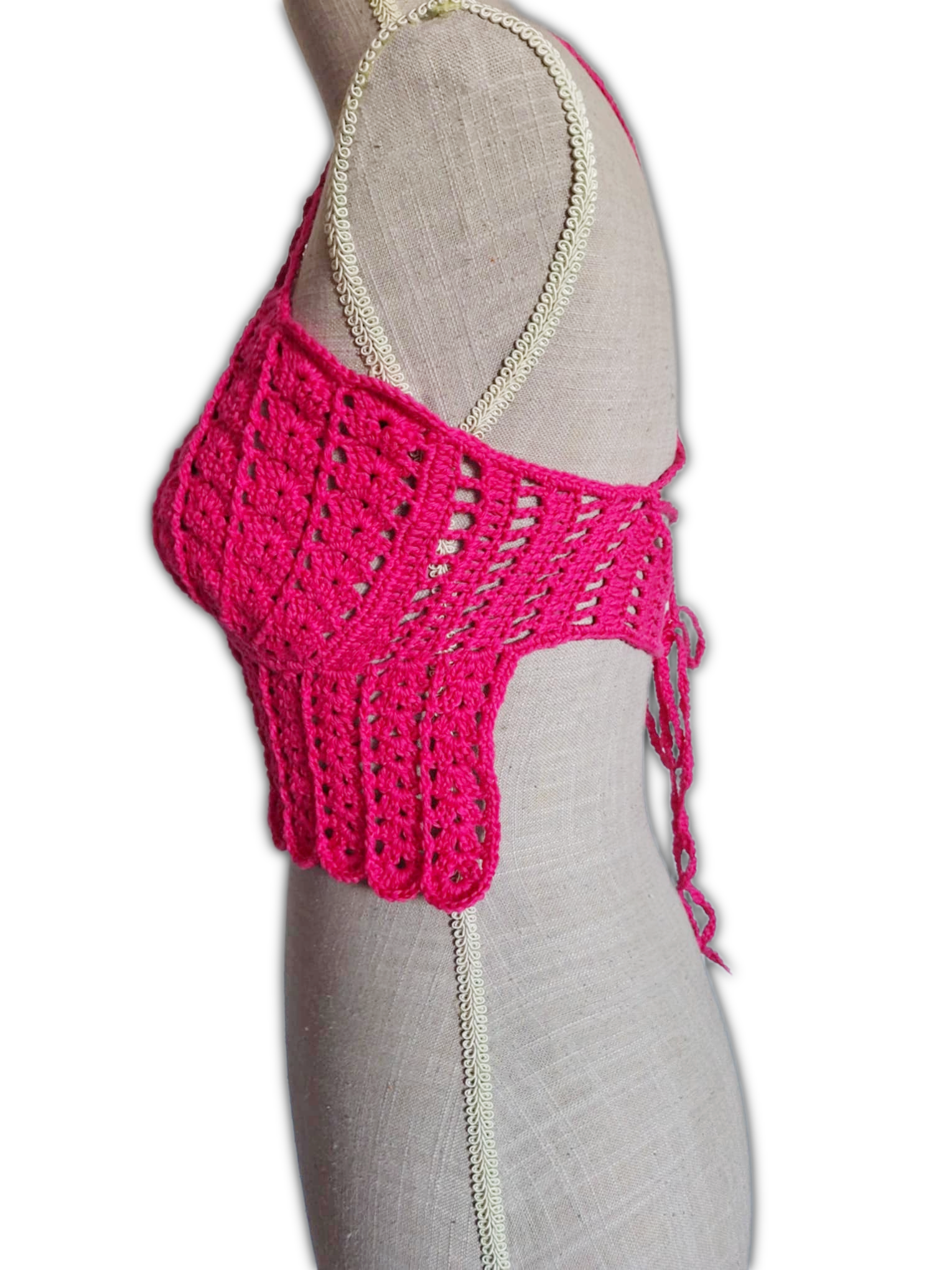 Vista lateral de um top de crochet fúcsia num manequim. A imagem mostra o perfil do busto com copas em forma de conchas, as alças finas sobre o ombro e parte da lateral aberta do top, destacando o caimento e a estrutura leve da peça.
